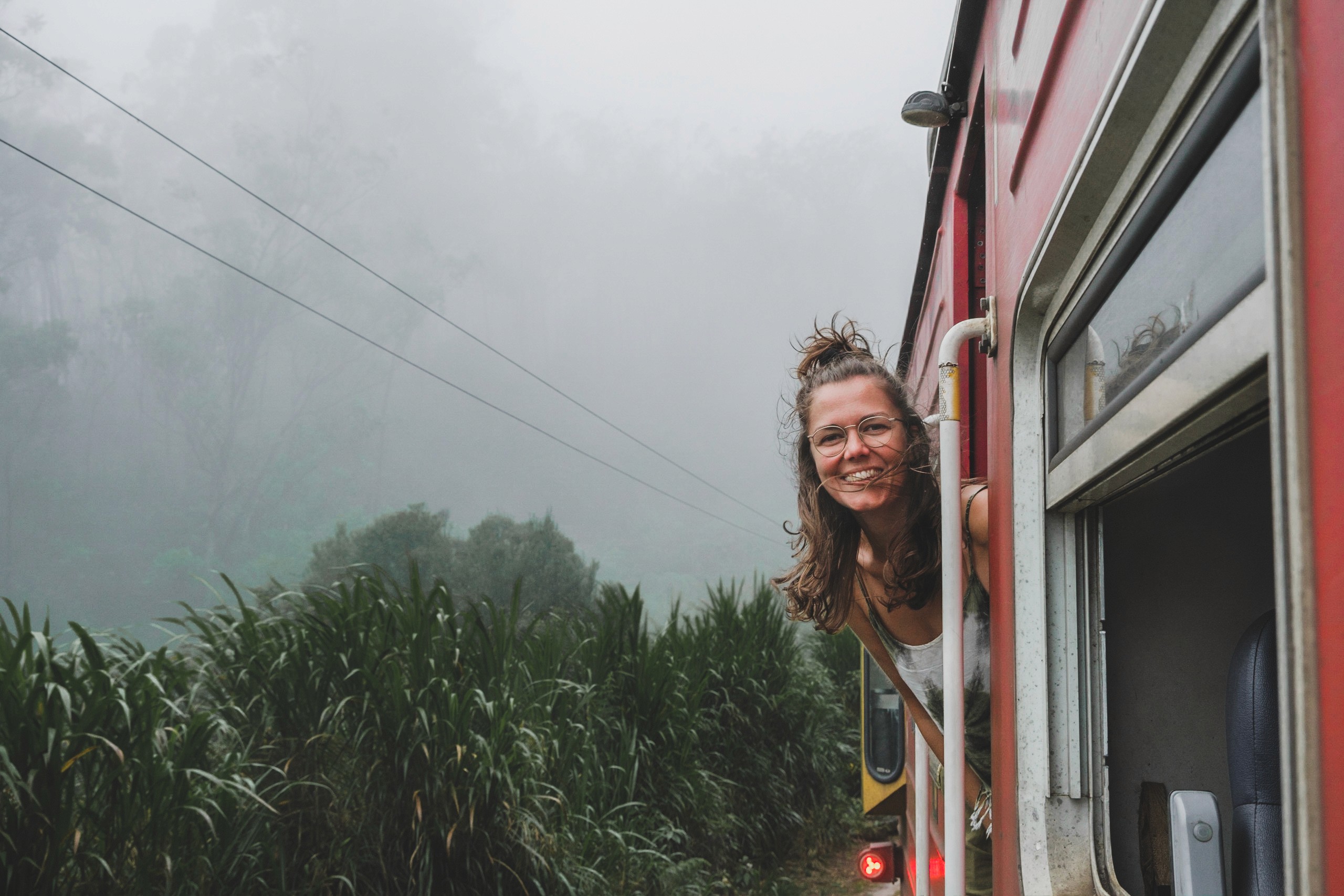 Eva leaning out of a train window surrounded by lush greenery and fog.
