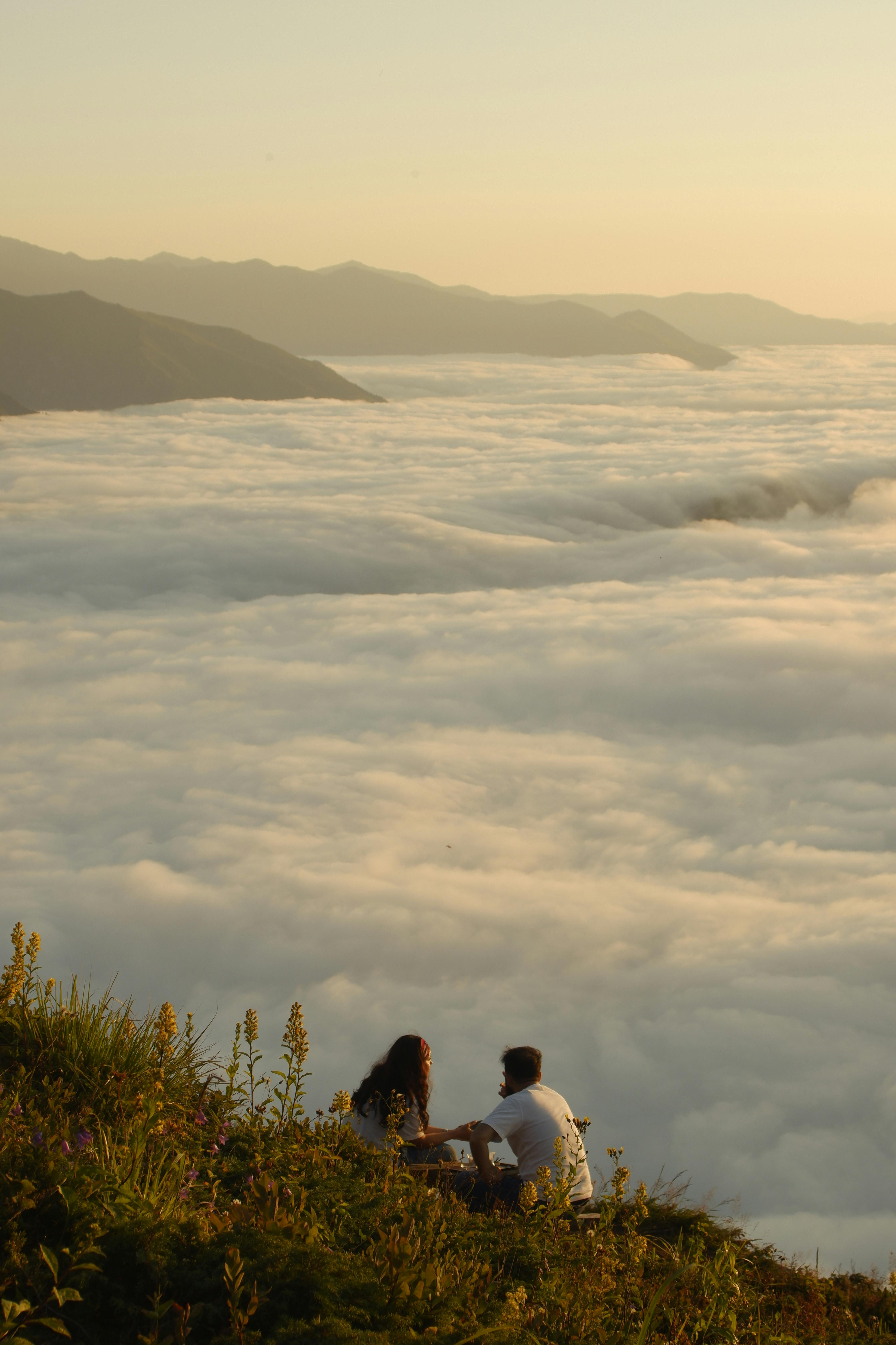 Two people sit on grassy ground overlooking a sea of clouds with mountains in the background during golden hour.