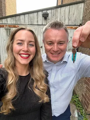 Couple smiling together holding house keys, woman with blonde hair and man in light blue shirt, standing in a garden