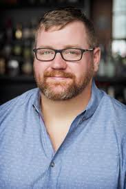 Josh, white man with glasses wearing a blue button down shirt and smiling at the camera
