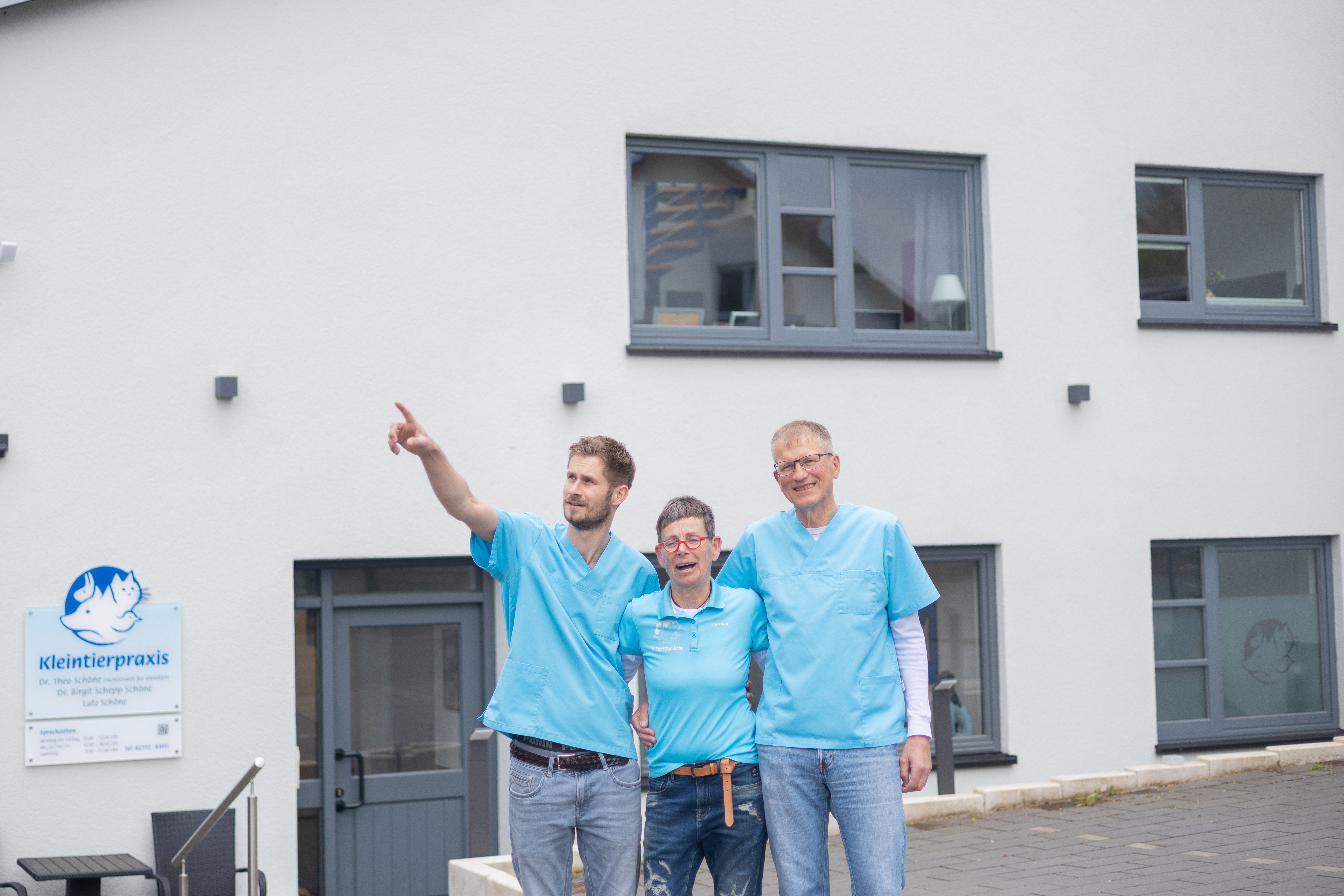 Drei lächelnde Tierärzte der Tierarztpraxis "Kleintierpraxis" stehen vor weißem Gebäude. Mann zeigt.