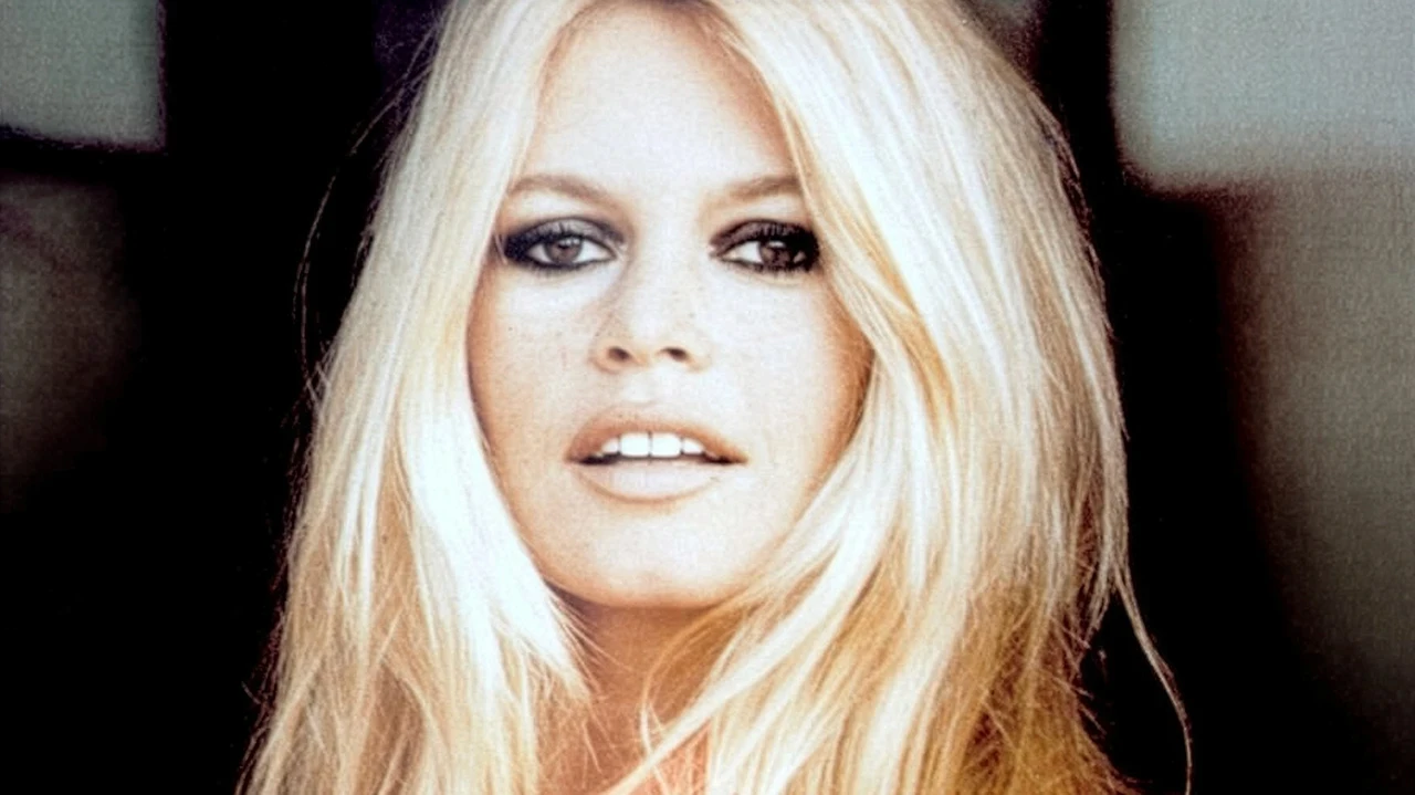 Portrait en gros plan d’une femme blonde aux cheveux longs, regard direct vers l’objectif, maquillage prononcé autour des yeux et arrière-plan sombre flou. L’image accompagne l’article “initials TT, comment opter pour une stratégie de télétravail efficace”, en suggérant une figure d’icône ou d’initiales symboliques.