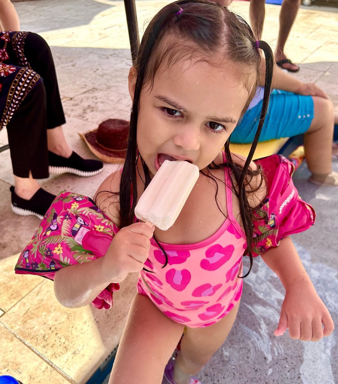 A little girl in a bright pink swimsuit biting into a white lime flavored popsicle