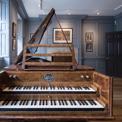 Un clavecín en una sala bien iluminada y elegante, con cuadros enmarcados en paredes grises y suelo de madera.