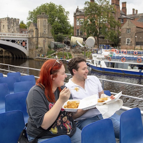 一名男子和一名女子坐在船上,享用外卖盒中的食物。背景中可以看到一座桥梁、几栋建筑物,以及另一艘船。