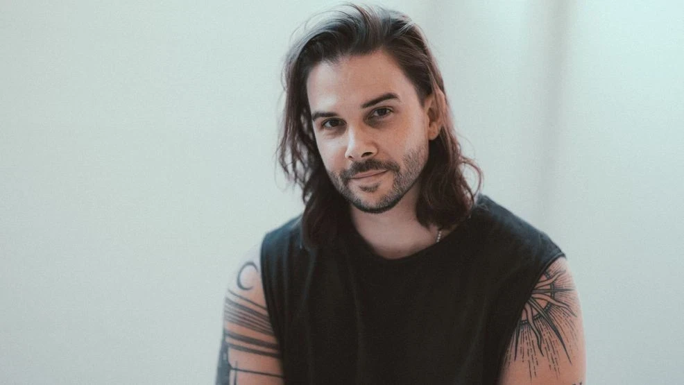 A man with long hair and tattoos sits against a light background, looking slightly serious.