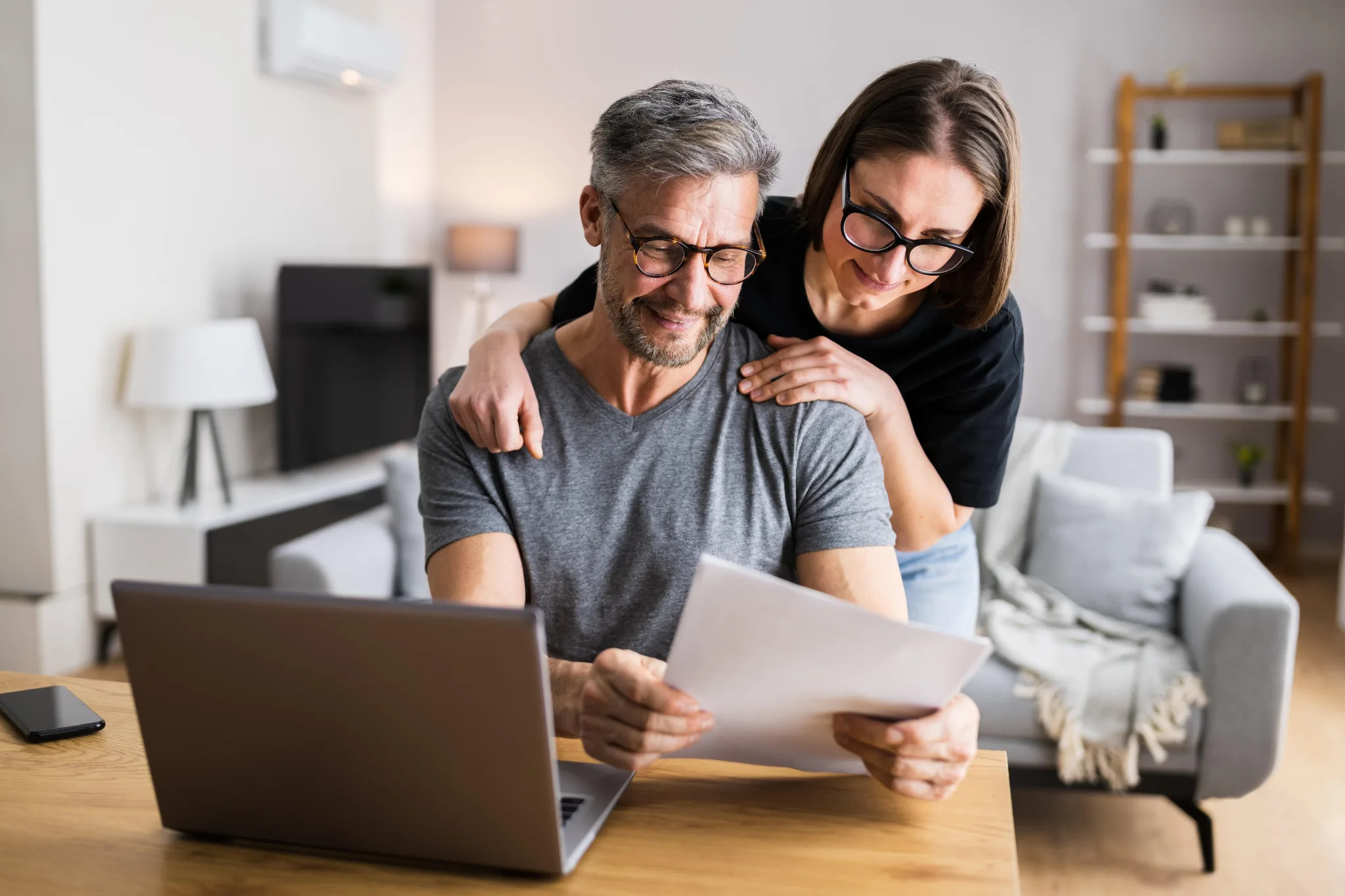 A man and woman wearing glasses smile while reviewing documents together at a wooden desk with a laptop in a modern living room.