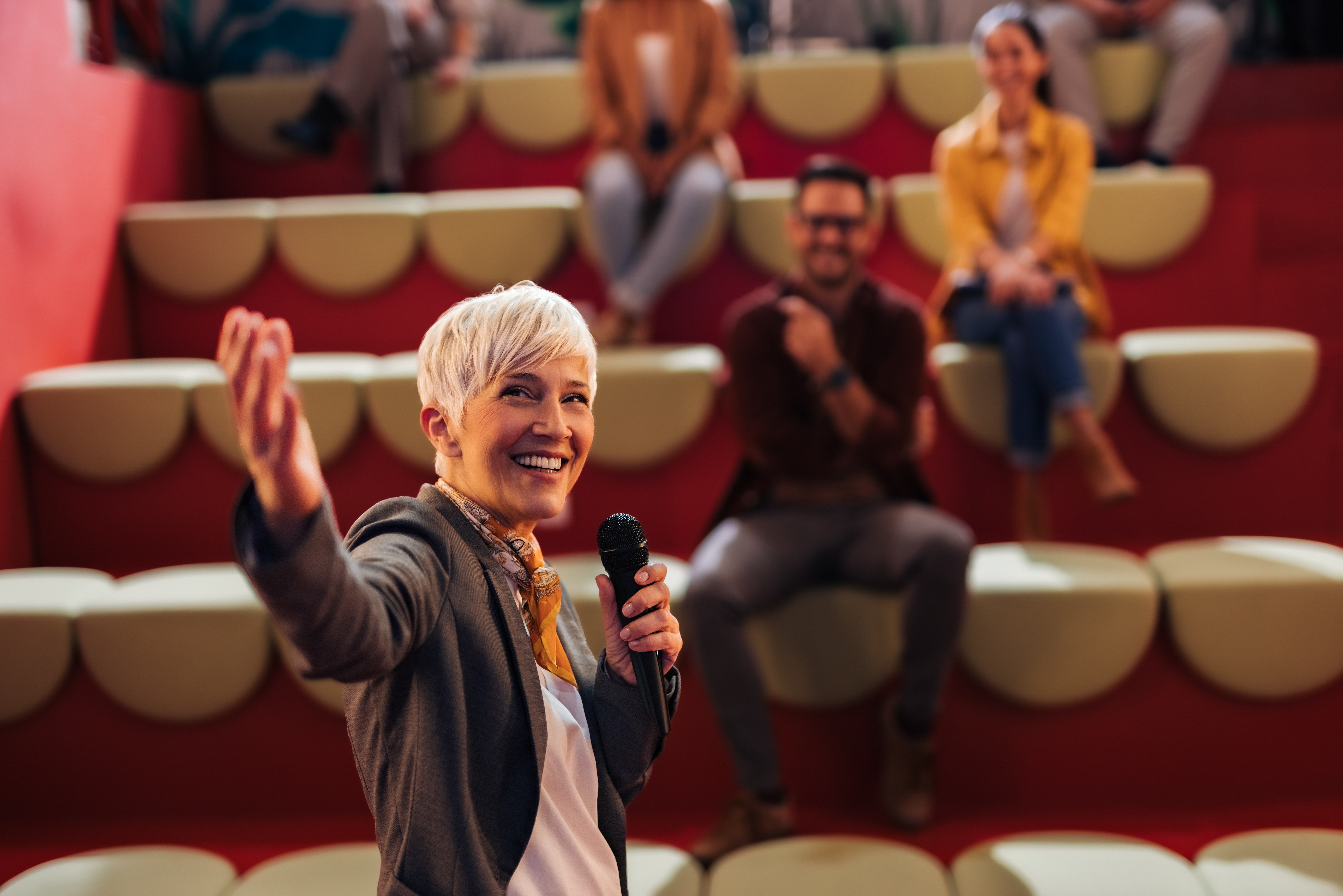 hero section background with plenty of space to the left out of focus; subject is a woman of color giving a speech from a podium with a smile and a laugh and confidence while others look on appreciatively