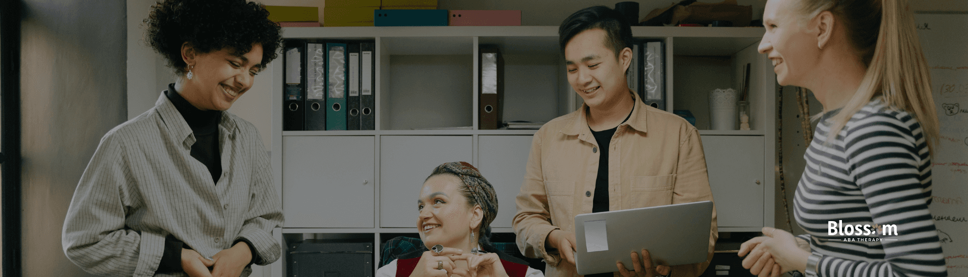 Diverse coworkers smiling and talking together in an office, representing teamwork and positive communication; Blossom ABA Therapy logo visible.