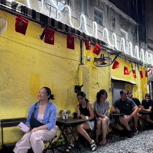 Folk sidder ved små caféborde udendørs mod en gul væg med hængende røde flag, lyskæder og en markise ovenover.
