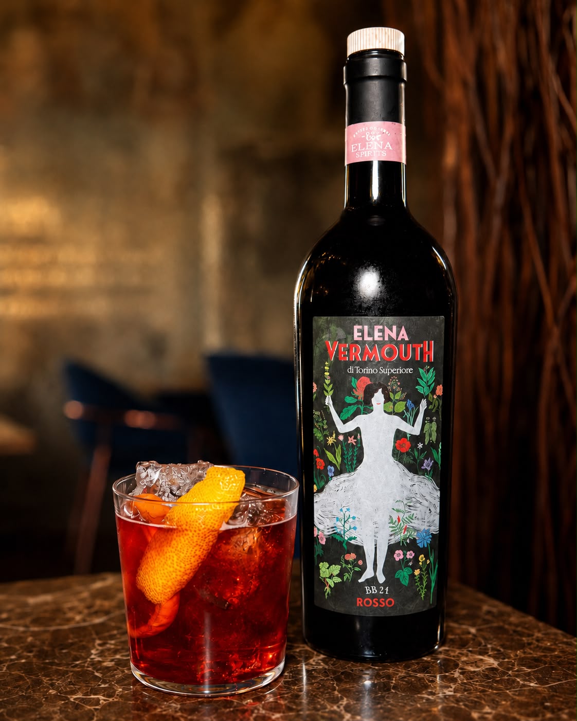 Cocktail rosso scuro in tumbler con fetta di arancia e bottiglia di vermouth Elena Superiore