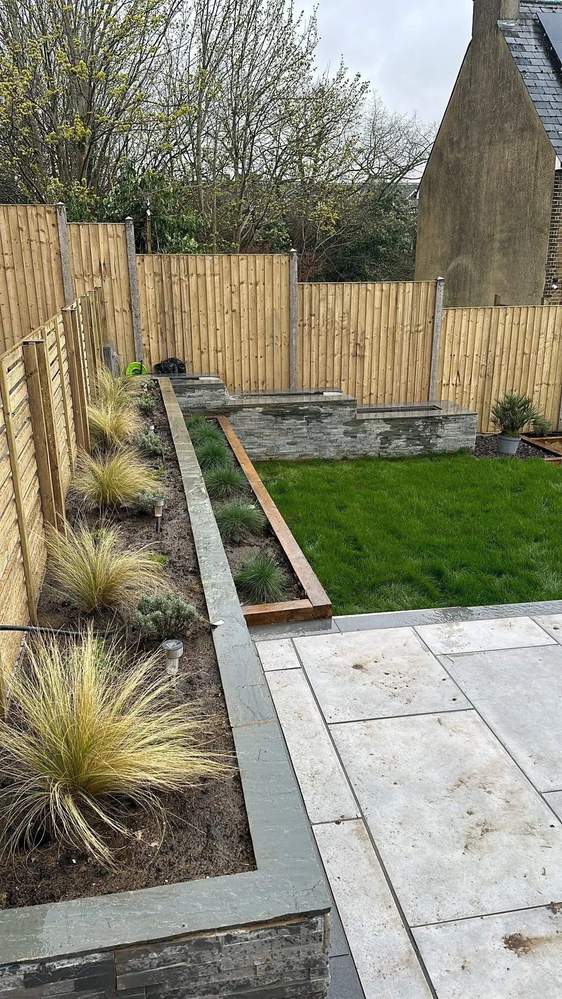 A landscaped garden with neatly arranged plants, a grassy area, and a paved pathway beside wooden fencing.
