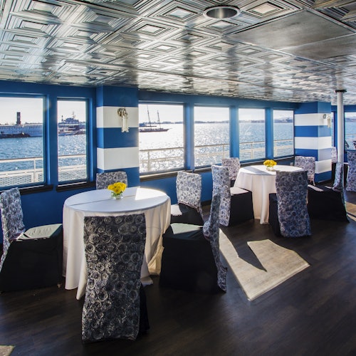 Una habitación con paredes rayadas, mesas redondas con manteles blancos y centros de mesa florales, y grandes ventanas con vista al puerto.