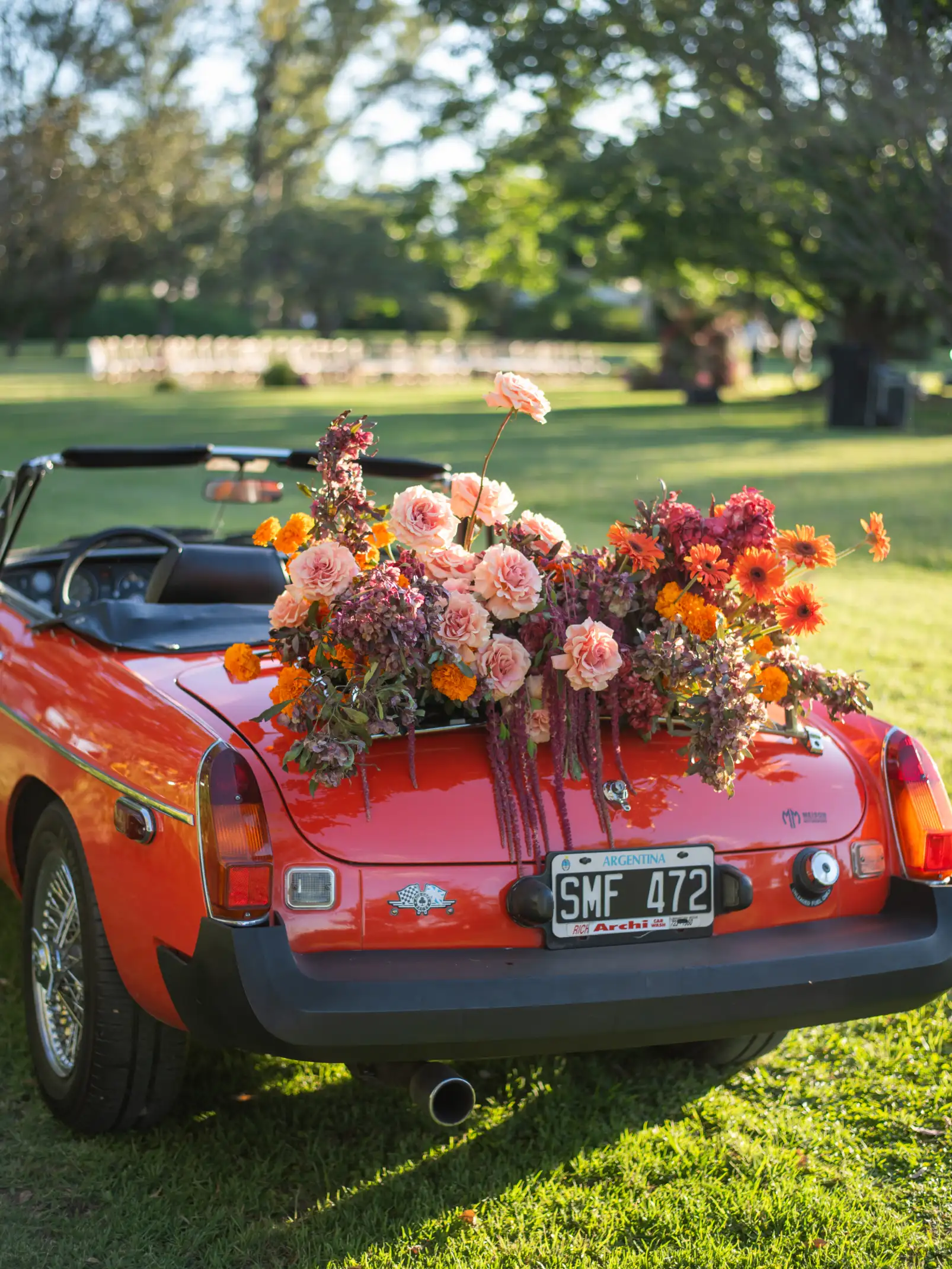 Auto clásico rojo decorado con flores rosadas y naranjas en la parte trasera, estacionado sobre césped en entorno natural.