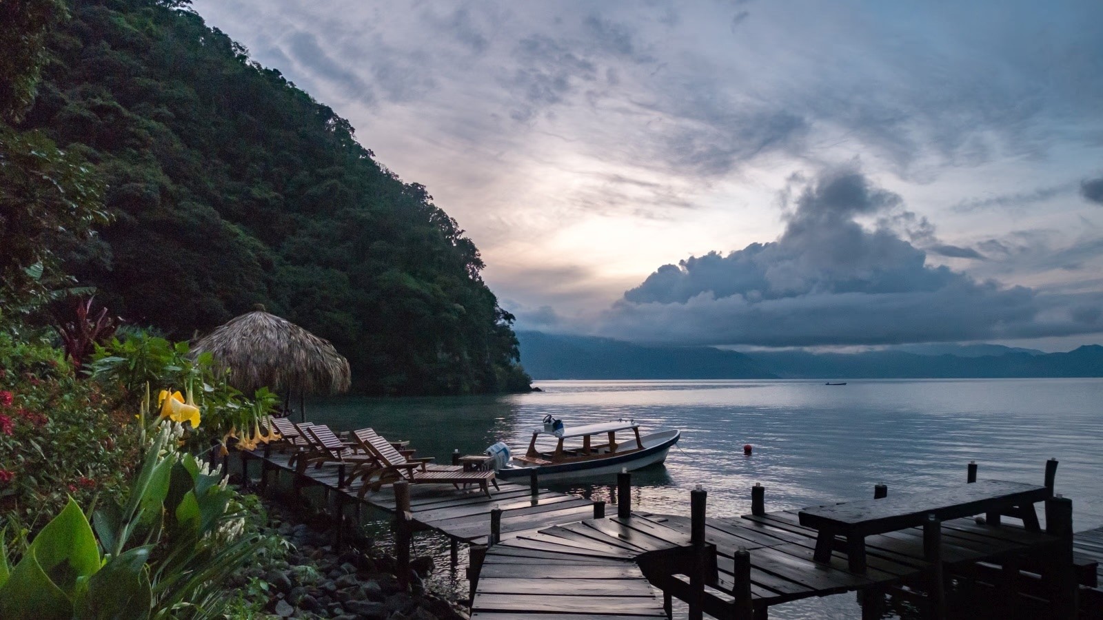 Un muelle de madera con sillas de playa y un pequeño bote en un lago tranquilo al atardecer, rodeado de una ladera verde y frondosa.