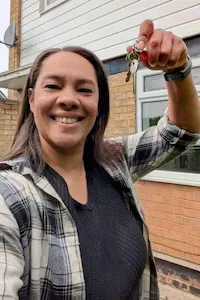 Woman in plaid flannel shirt smiling and holding up house keys in front of a residential property