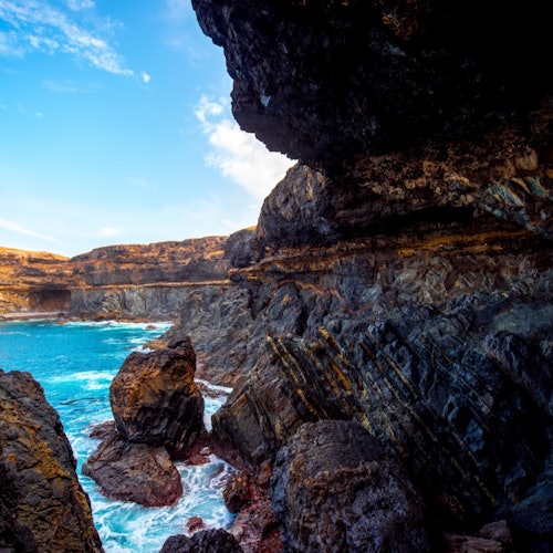 崎岖的海岸悬崖上布满黑色火山岩地貌，湛蓝的海水在部分多云的蓝天下闪耀。