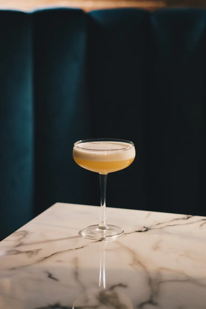 A classic coupe glass filled with a creamy yellow cocktail sits elegantly on a marbled table against a dark, plush background, capturing a sophisticated bar ambiance.