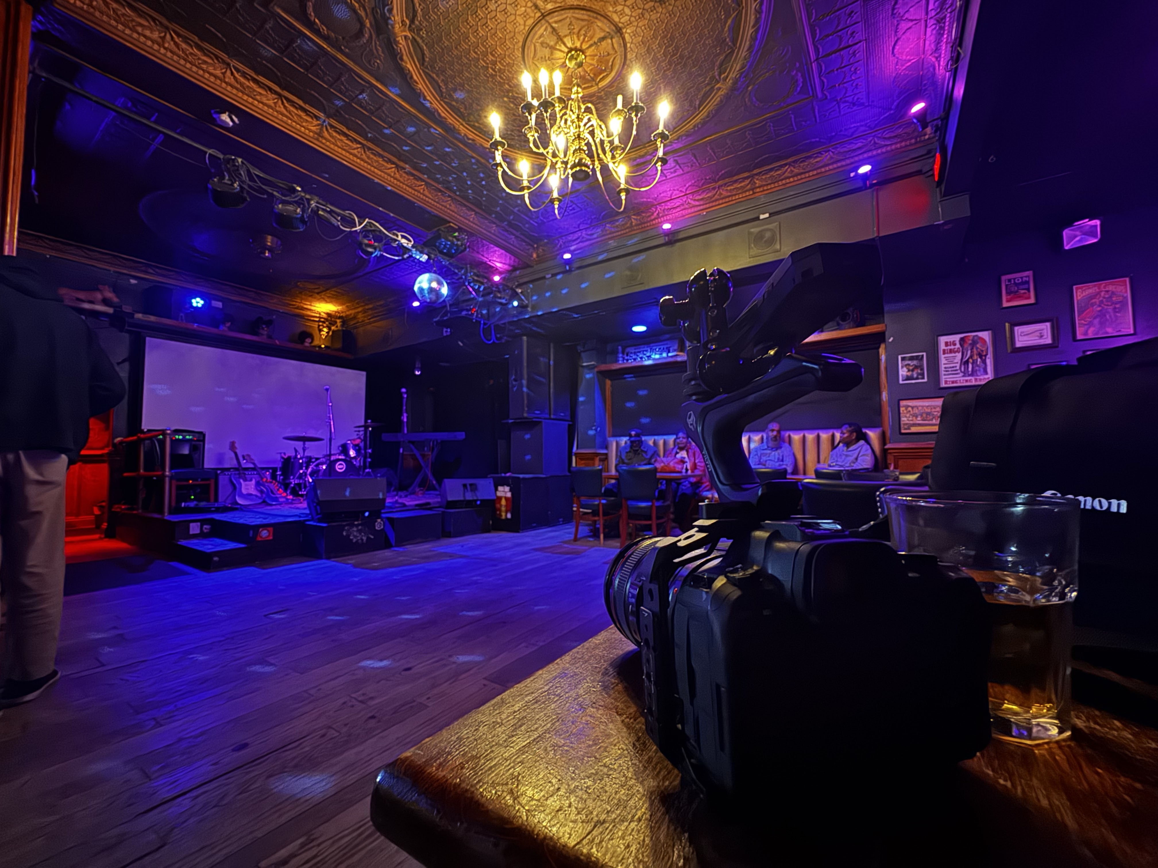 Behind-the-scenes view of a video camera framing a dimly lit bar stage with purple and blue performance lighting.
