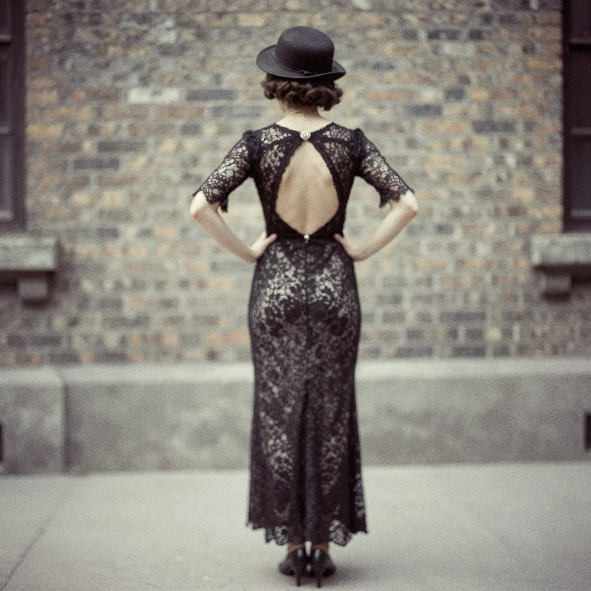 Woman in a black lace dress with an open back and bowler hat, seen from behind.
