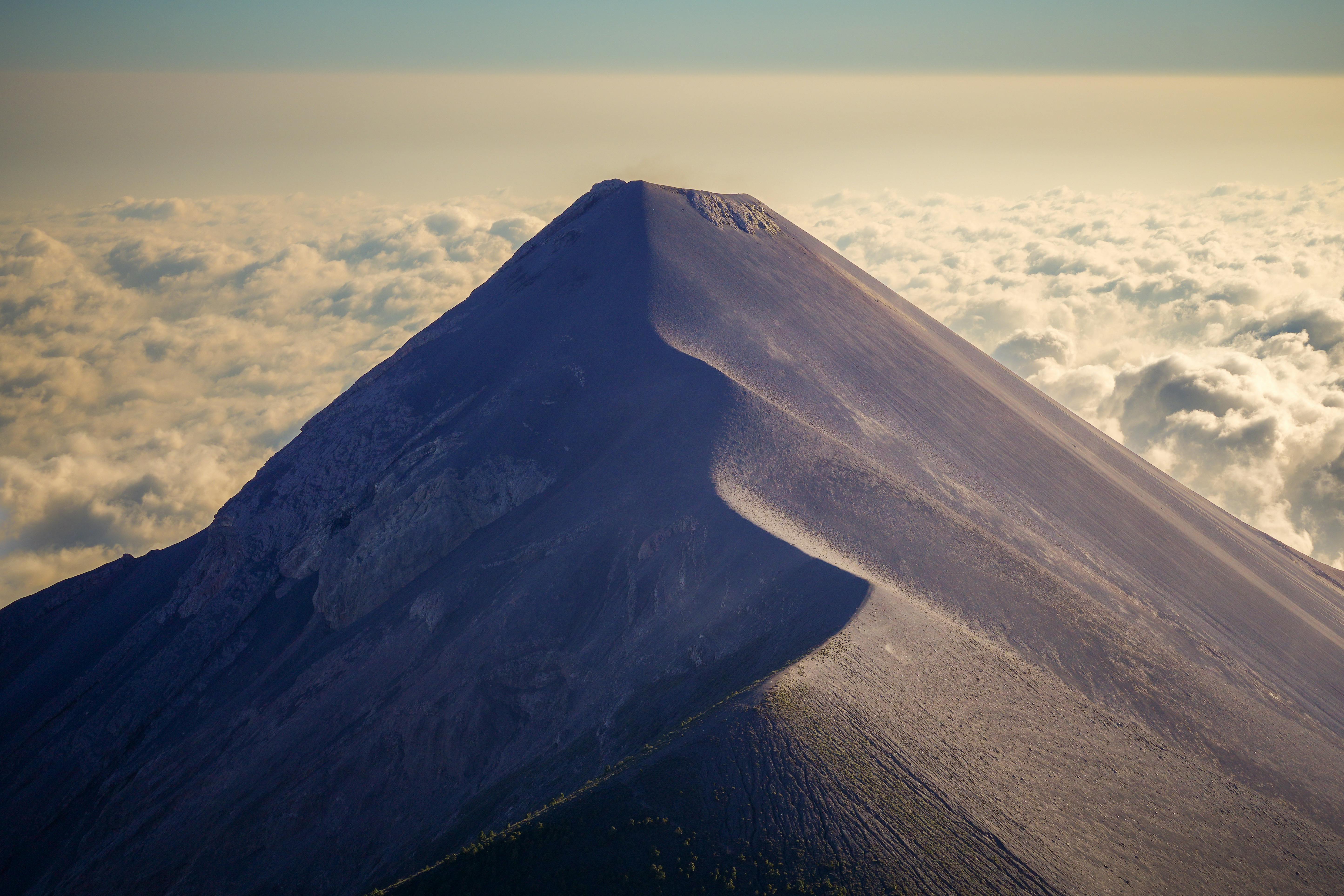 A majestic volcanic mountain peak rises sharply above a sea of fluffy, white clouds under a clear blue sky, showcasing its rugged, rocky surface.