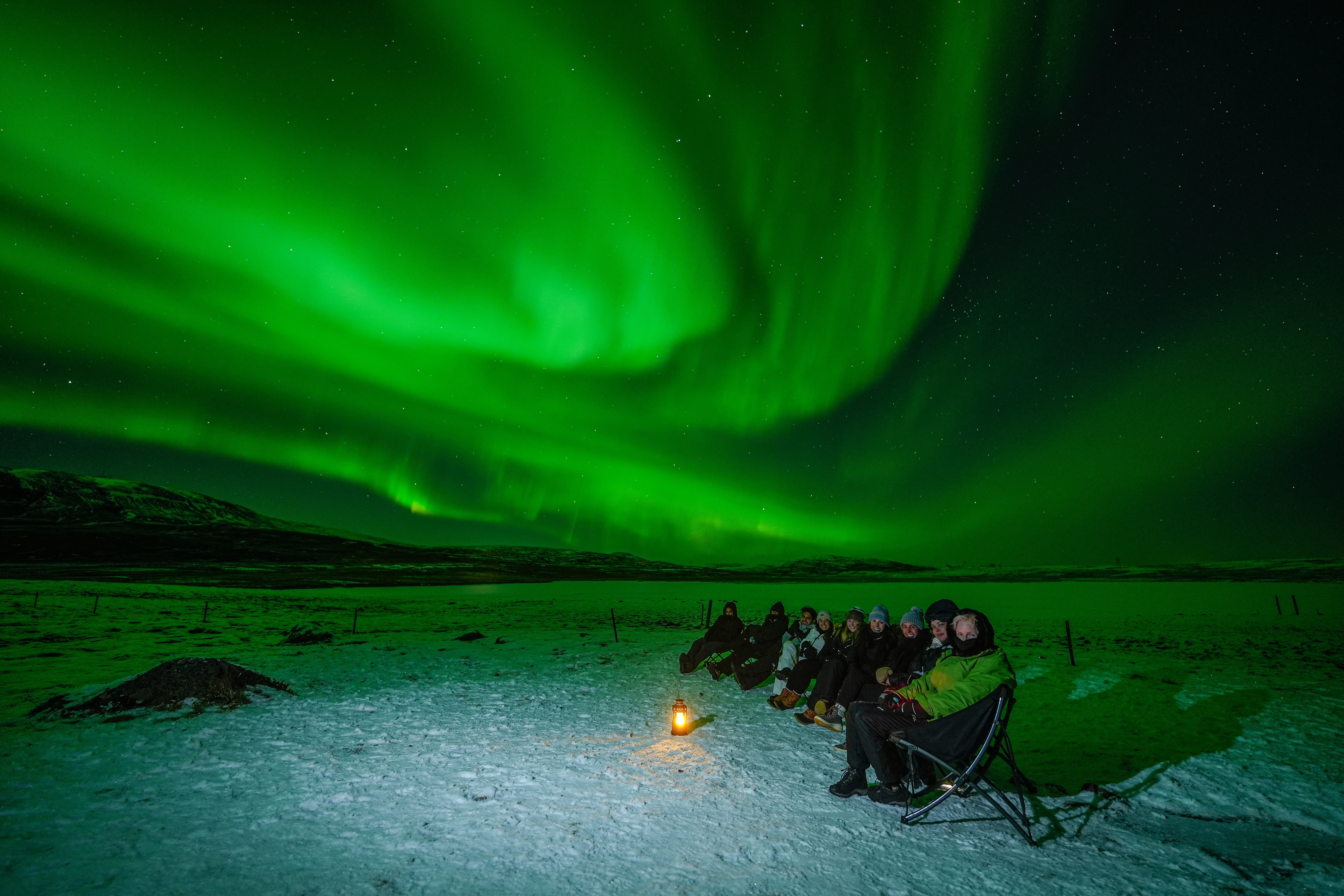 Small group sitting in camp chairs around a lantern beneath the northern lights