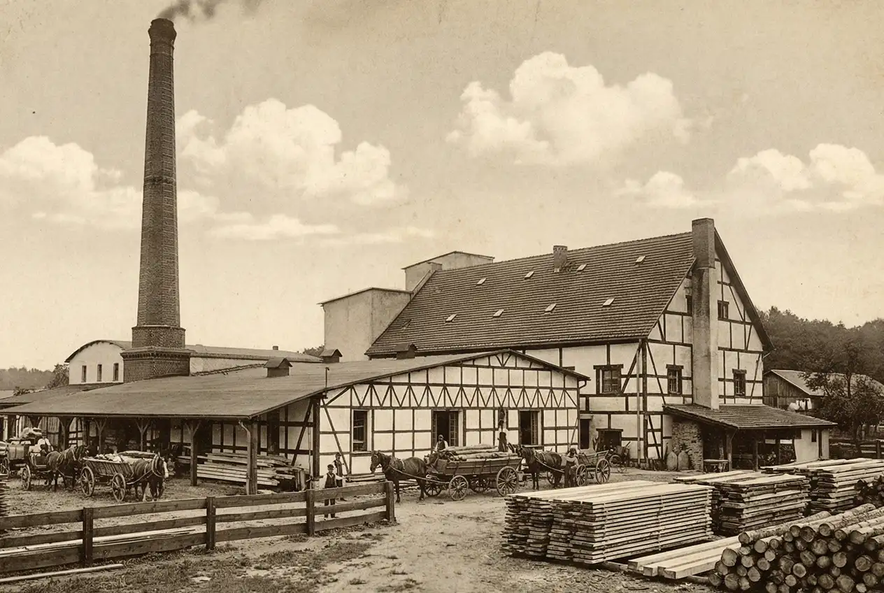 Stare zdjęcie tartaku z wysokim kominem i halami produkcyjnymi, przedstawiające historyczny wygląd obiektu.