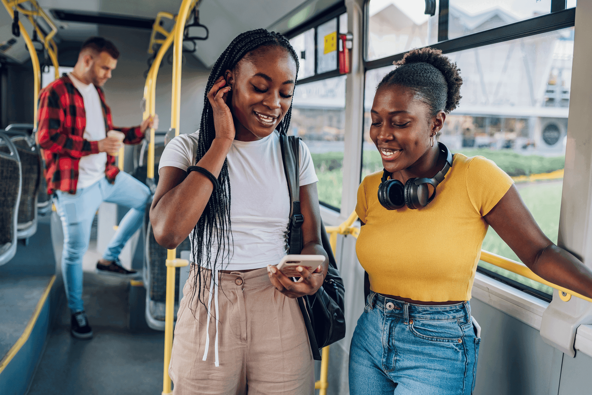 Two women smiling and looking at a smartphone while standing on a bus, with one wearing headphones around her neck and the other wearing a white t-shirt and tan pants, capturing a moment of modern connectivity and public transportation.