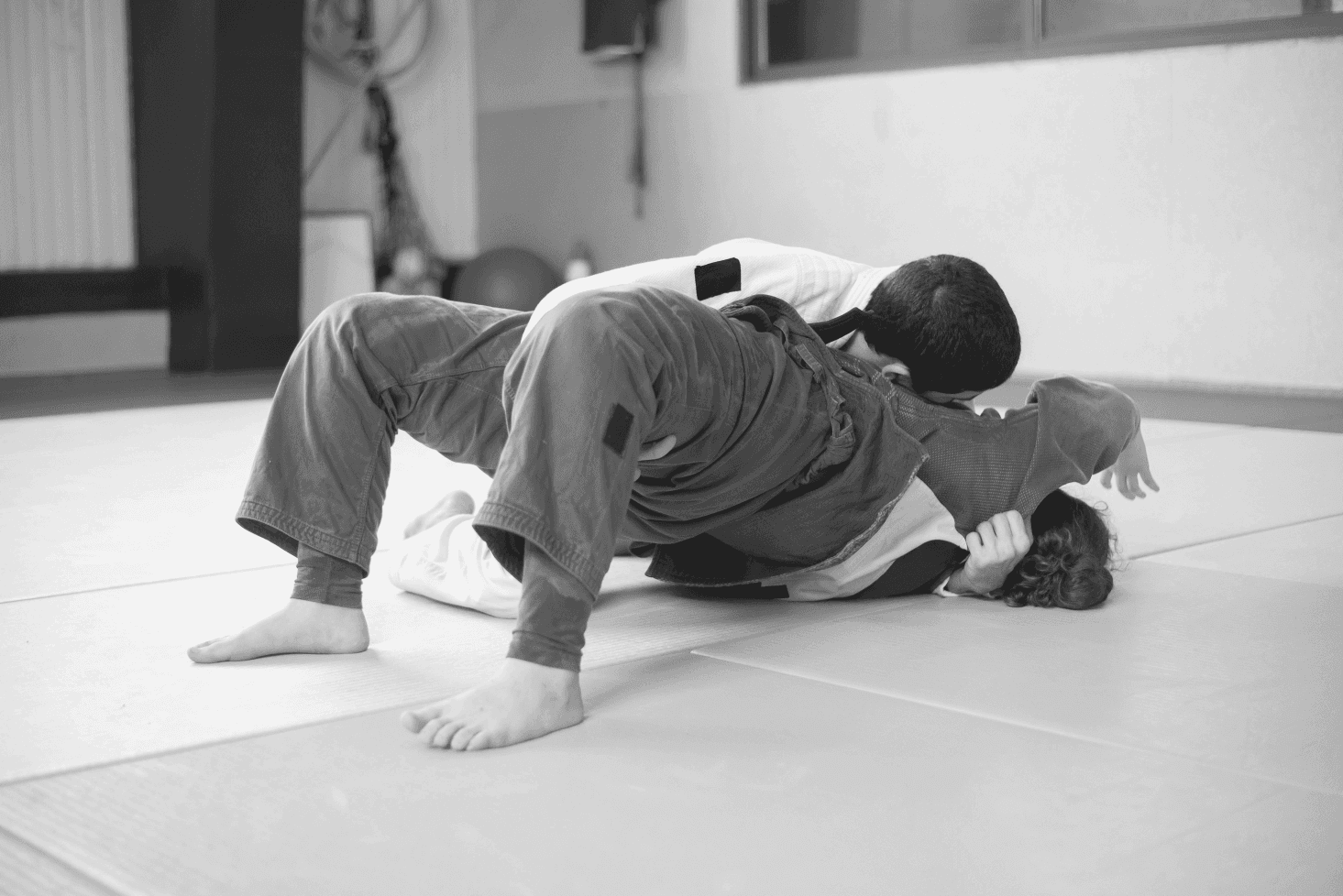 Two young students grappling on a yellow mat, with one student in a blue gi maintaining a top position over a training partner wearing a white gi.