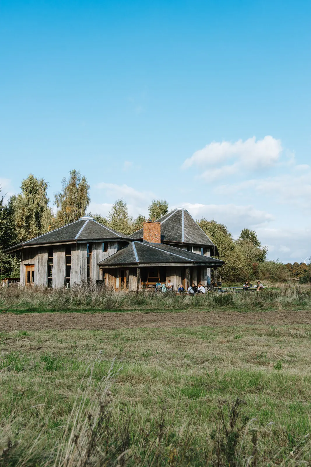 Een houten gebouw met puntdaken achter een grasveld onder een blauwe lucht, op het terras zitten mensen