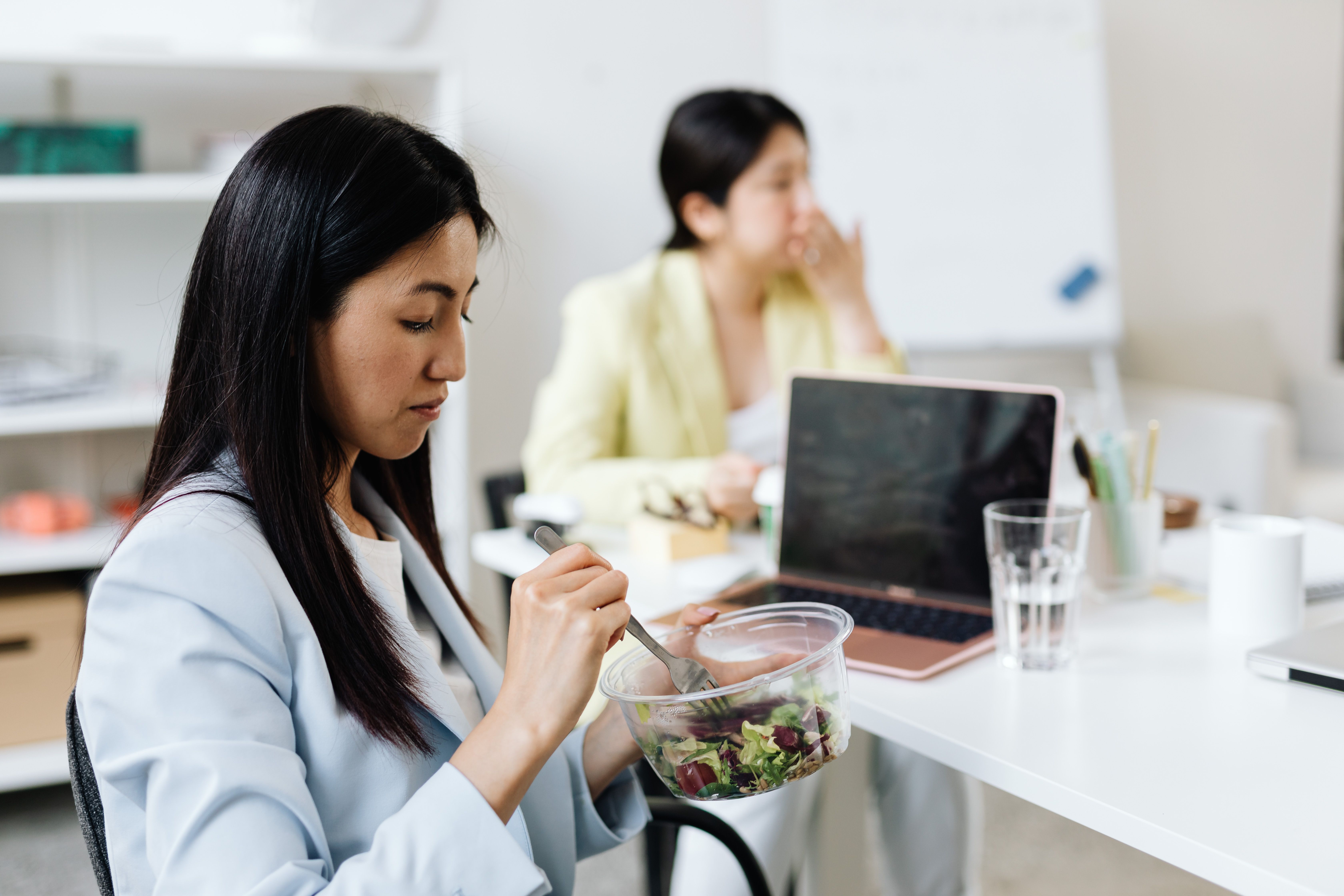 Woman in a blue blazer eating a salad at her office desk