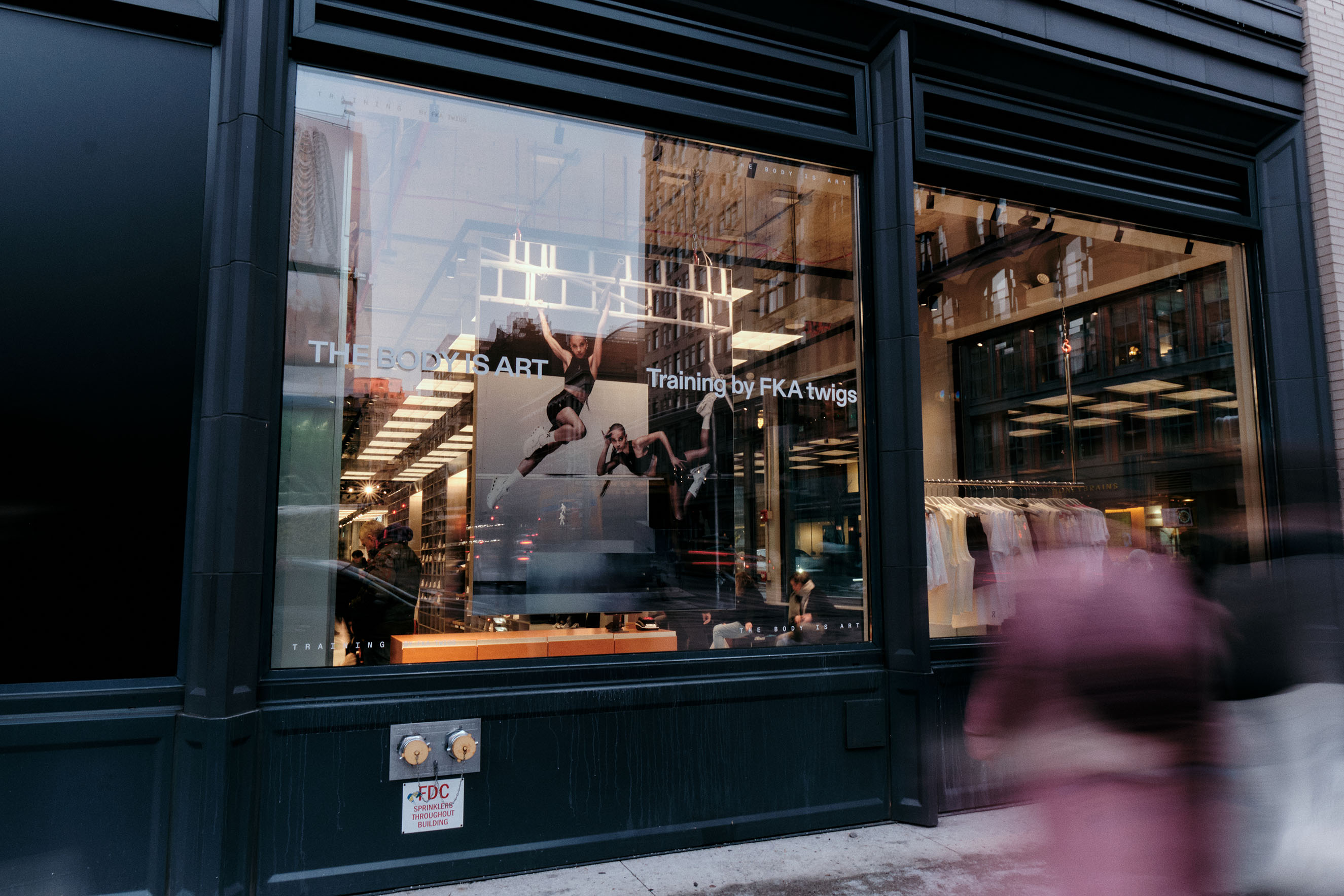A storefront window displays striking visuals: a large poster reading THE BODY AS ART Training by FKA twigs, featuring dancers in mid-air. Blurred pedestrians walk by as the campaign draws eyes to clothing inside the store.