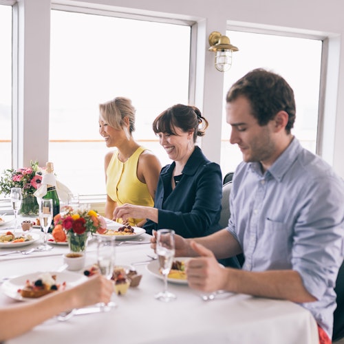 Cuatro personas cenando juntas en una mesa con flores y platos de comida en un barco o junto a grandes ventanas.