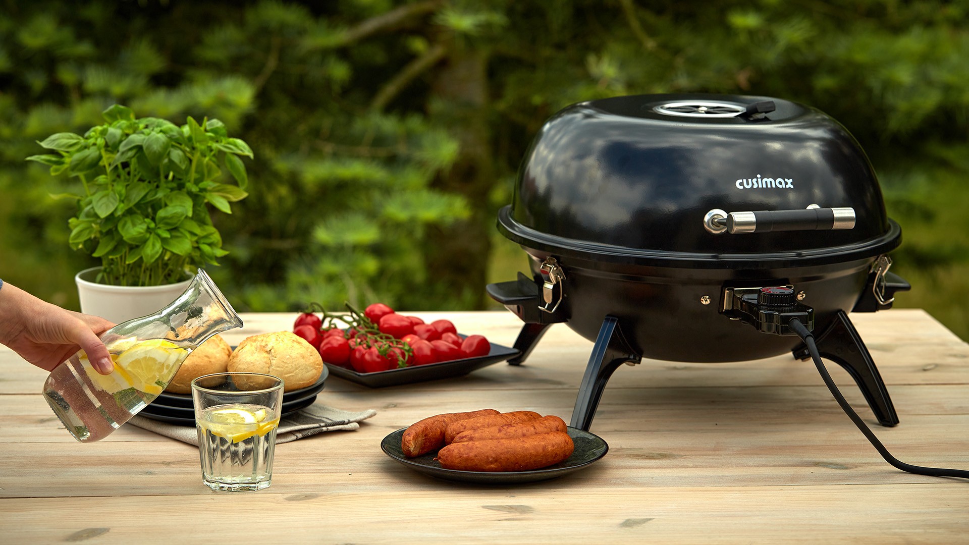 A portable black grill is set on a wooden table outdoors, surrounded by a glass of lemon water, a plate of buns, a dish of red cherry tomatoes, and a plate with breaded meat, with a vibrant basil plant in the background.