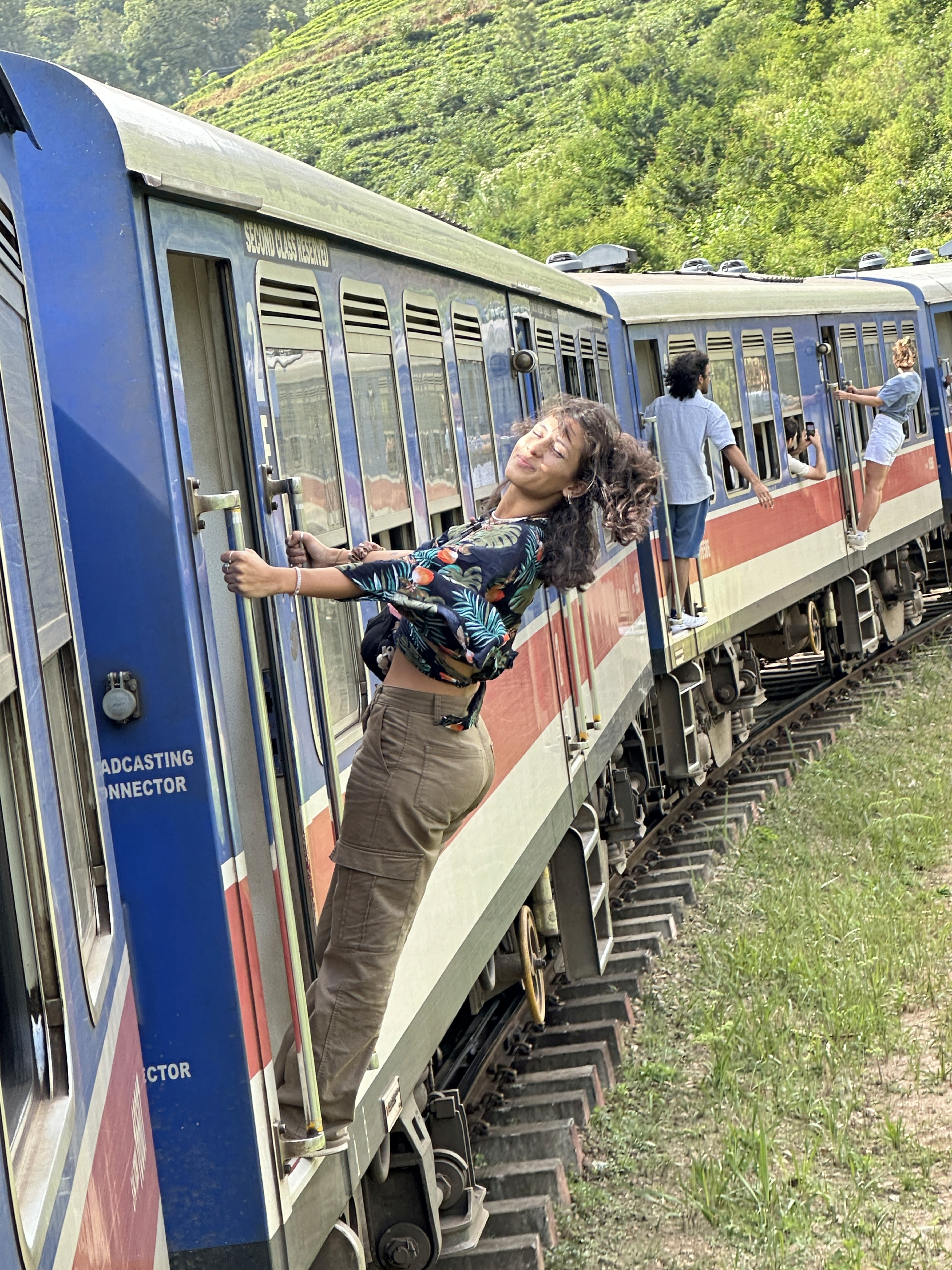 A carefree woman is hanging from the door of a slow-moving train, leaning back and smiling at the camera