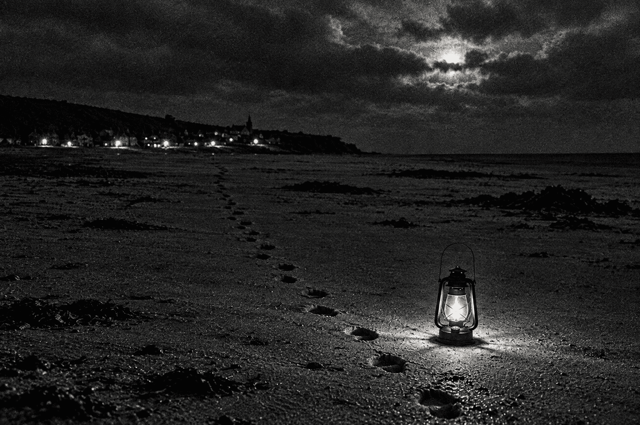 At night, a lit oil lantern on the empty seabed with footprints leading back to the village lights
