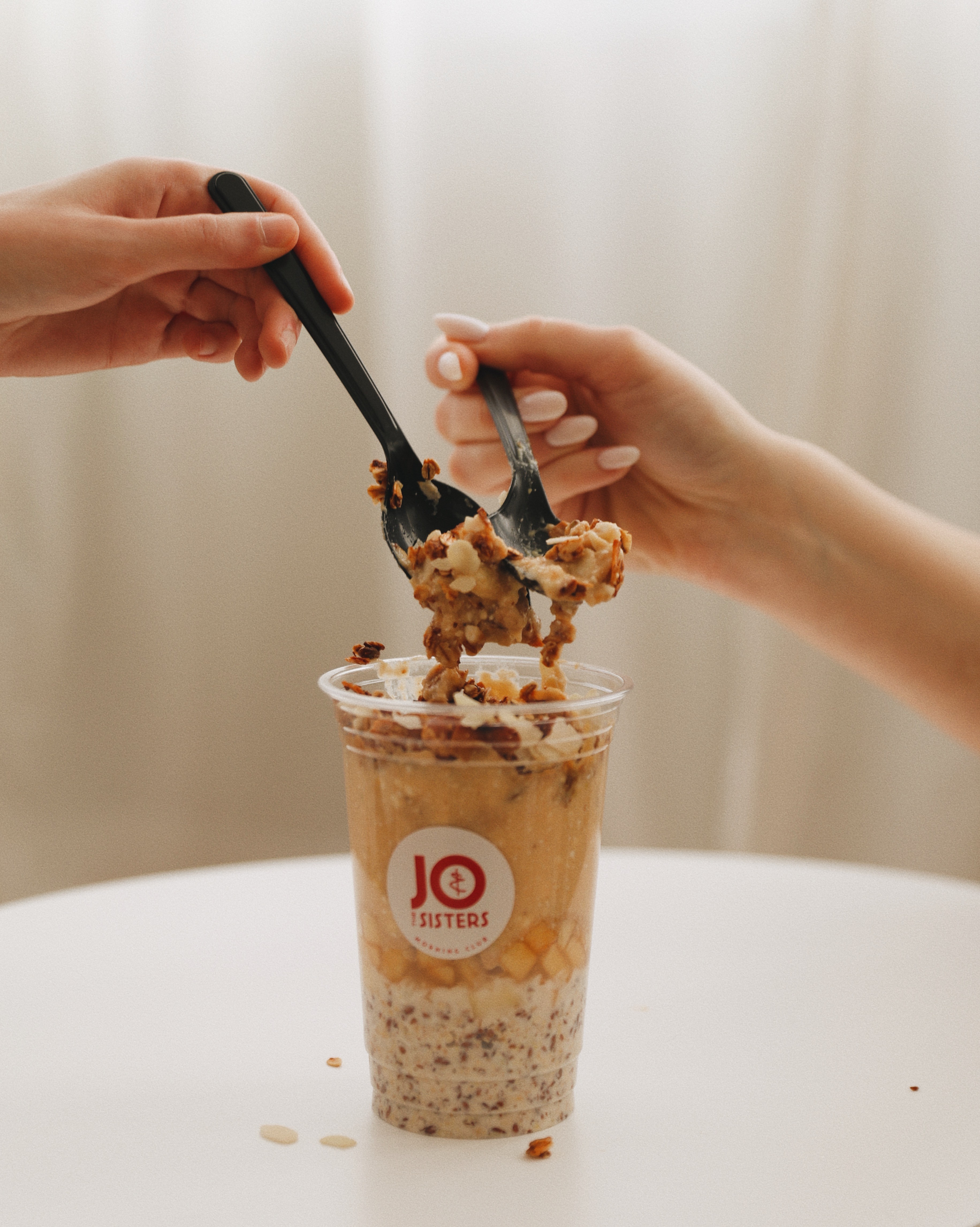 Two hands holding spoons scoop a layered breakfast cup with oats, fruit, and granola from Jo & The Sisters, highlighting a healthy, balanced meal.