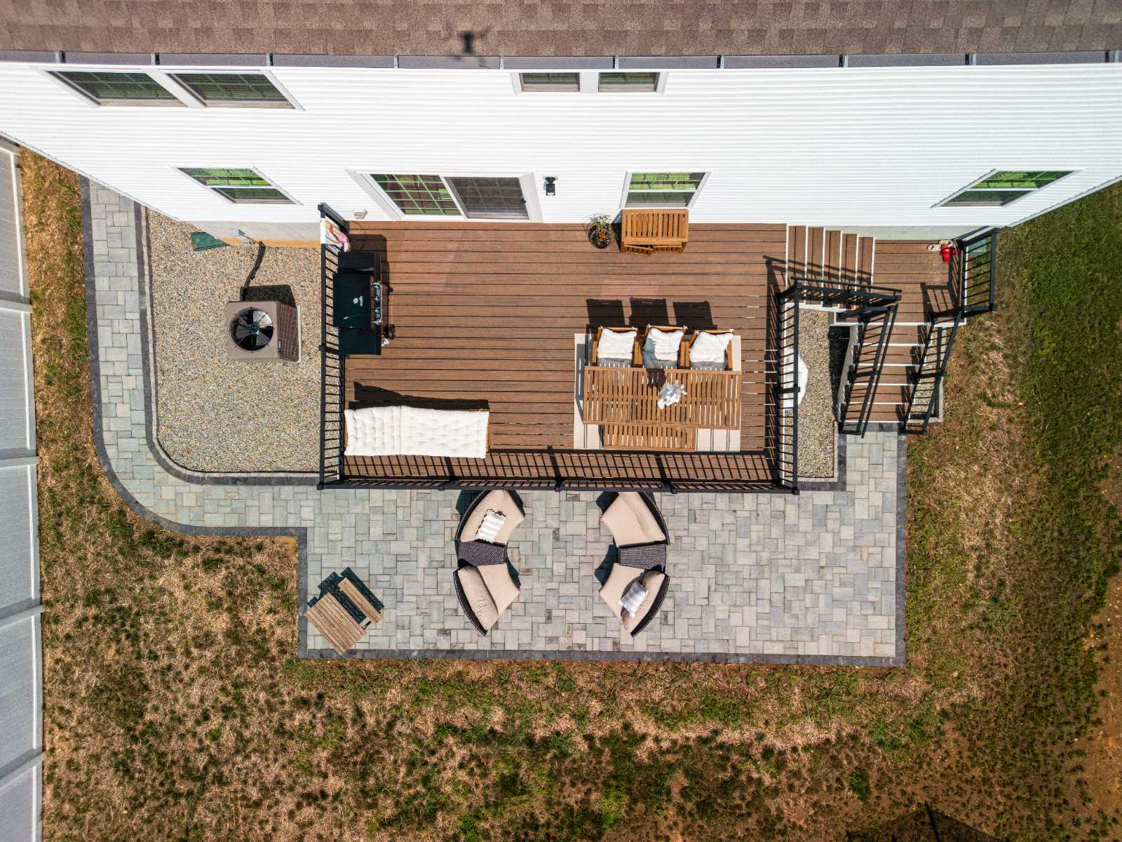A neatly arranged stone patio with a mix of beige and gray tiles is bordered by lush green grass, leading up to a white porch with steps and striped chairs, creating a welcoming outdoor space.