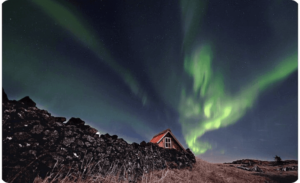 Green northern lights swirling above a small red‑roofed turf house behind a dark lava wall