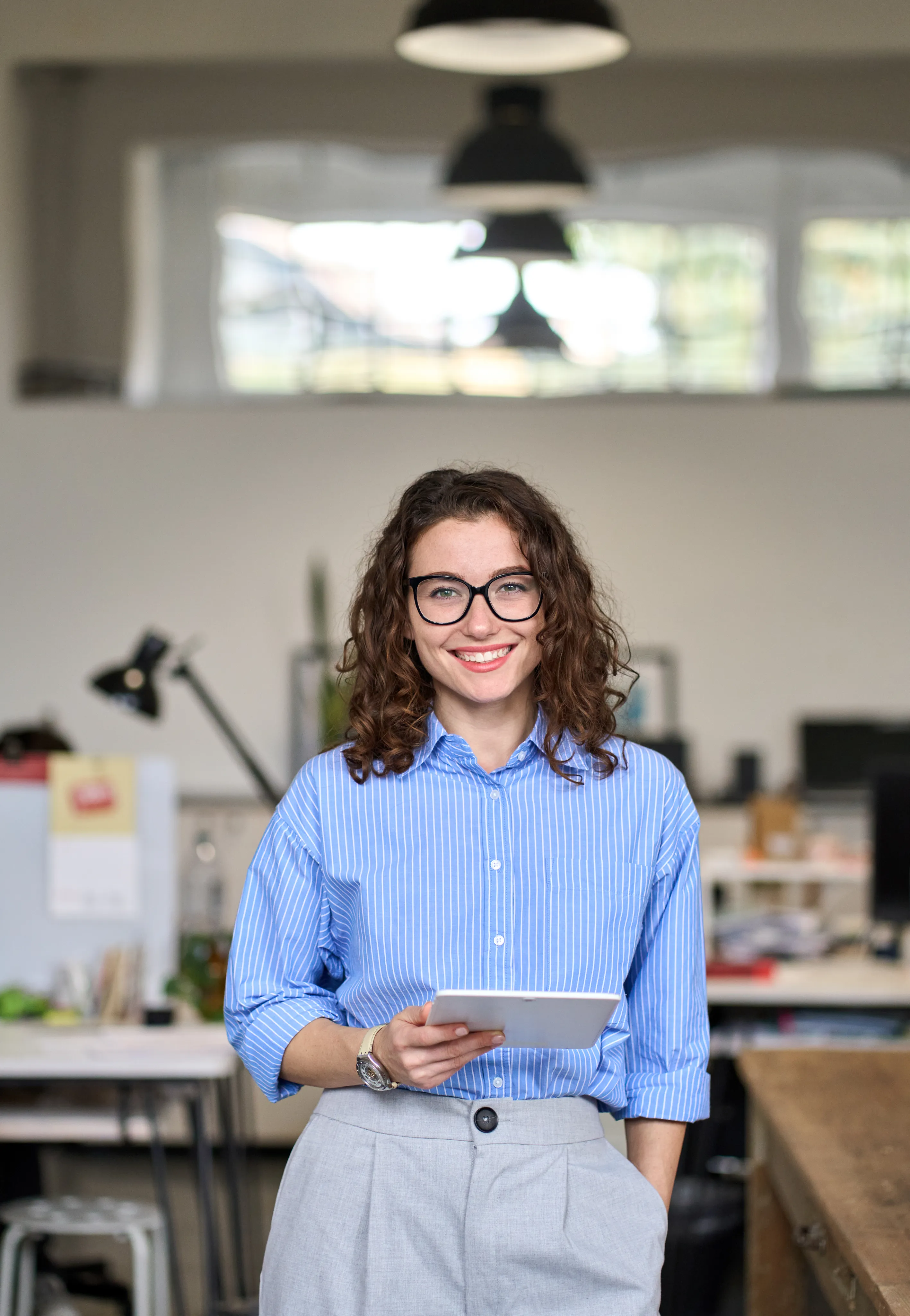 A smiling young woman with curly hair and glasses holding a digital tablet while standing in a bright, modern office space.