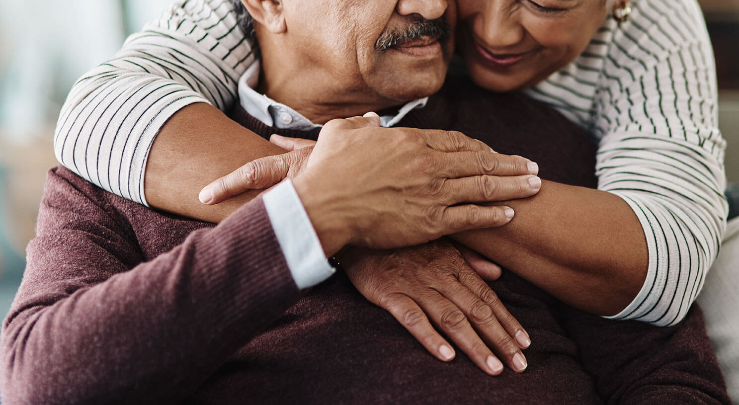 Close-up of a warm embrace between two older adults, with hands tenderly clasped. One person wears a burgundy sweater and the other a striped shirt.