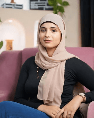 A young man with light brown hair is wearing a black suit jacket and white shirt, standing in a softly lit office setting, conveying professionalism.