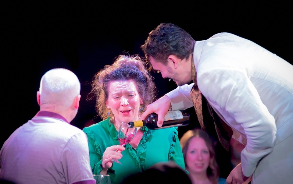 A Frow Show performer playing a waiter pouring wine for a guest