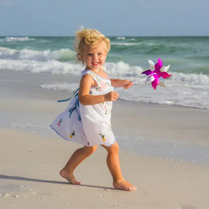 A young child in a white dress joyfully runs along a sunny beach while holding a colorful pinwheel, with gentle ocean waves in the background, capturing a playful moment in Bradenton, FL.