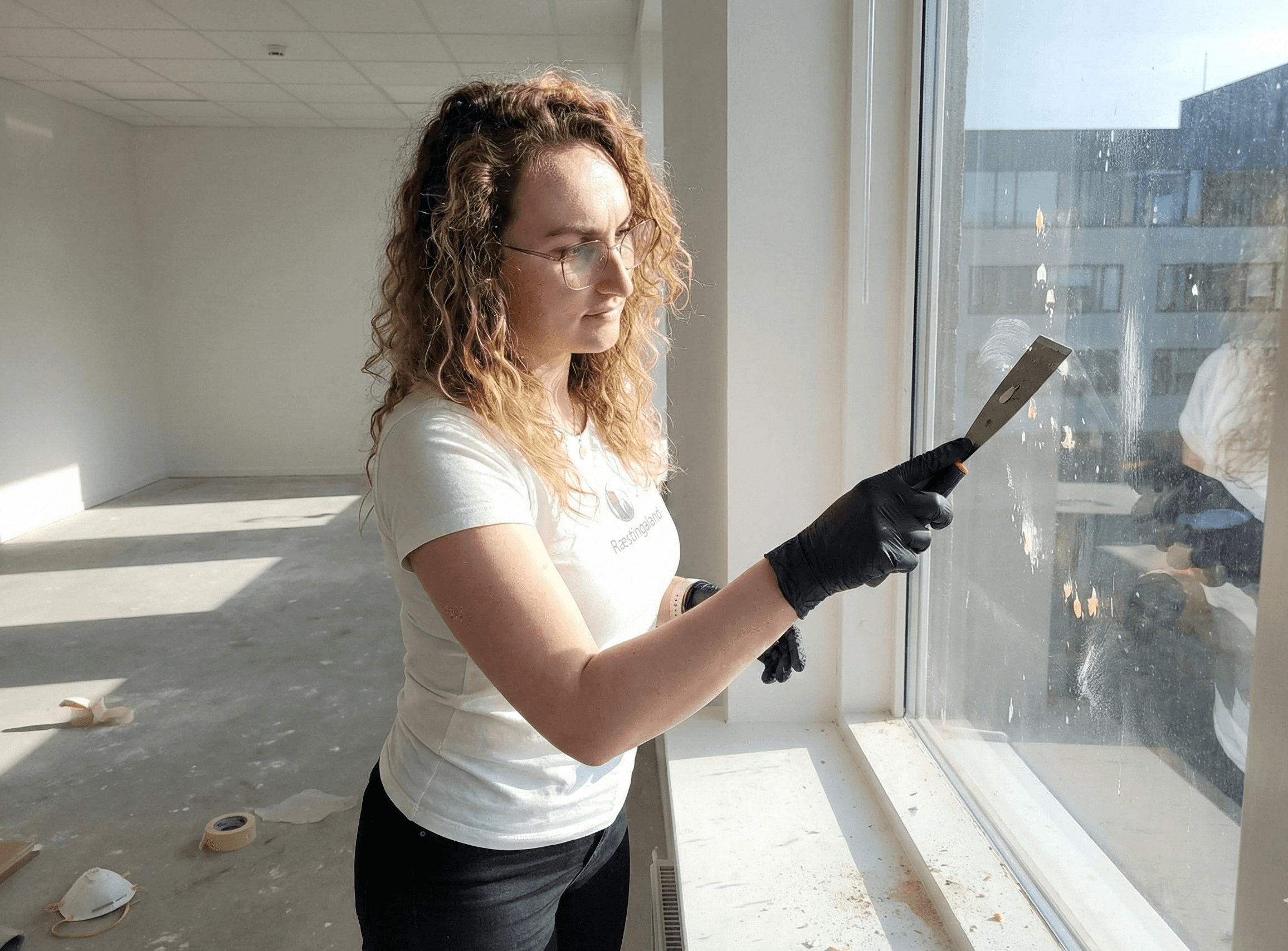 Fjarlæging á málningarslettum og lími af glugga með sköfu.