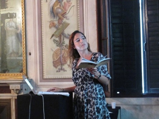 A photo of a person, with a book in her hand and giving a lecture while sitting in a room with frescoes on the walls.