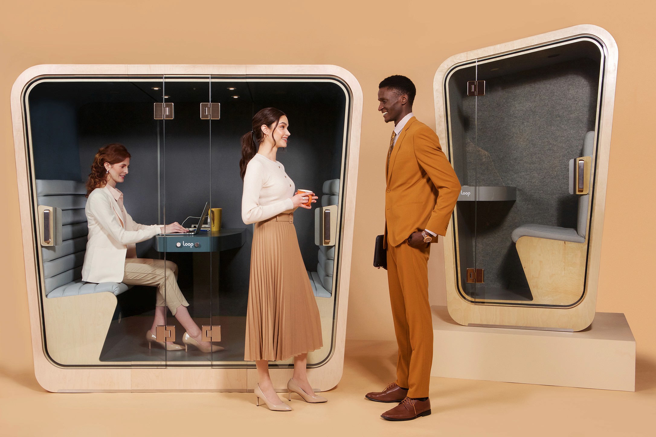 a man and a woman stand talking outside a privacy pod while another woman works on her computer inside the privacy pod