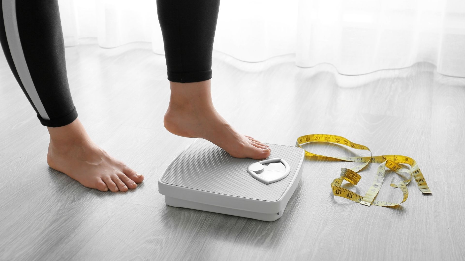 A person stands on a bathroom scale, with a measuring tape lying nearby on the floor.