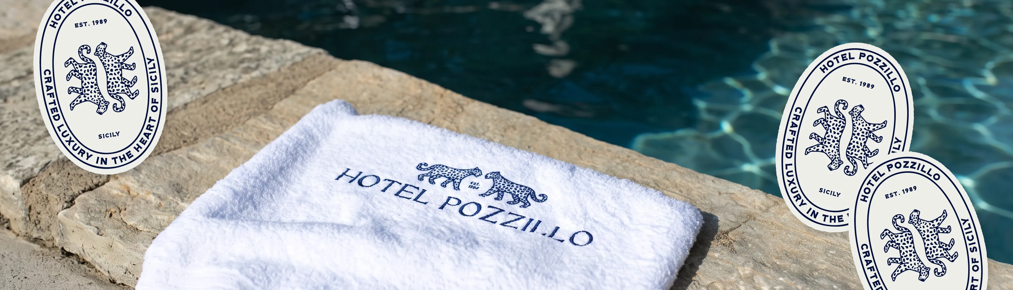 A white beach towel, with the Hotel Pozzillo logo embroidered on in blue thread, next to a pool