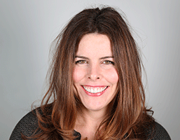 A woman with long, brown hair smiles at the camera against a light gray background. She wears a dark gray top and looks directly at the camera.